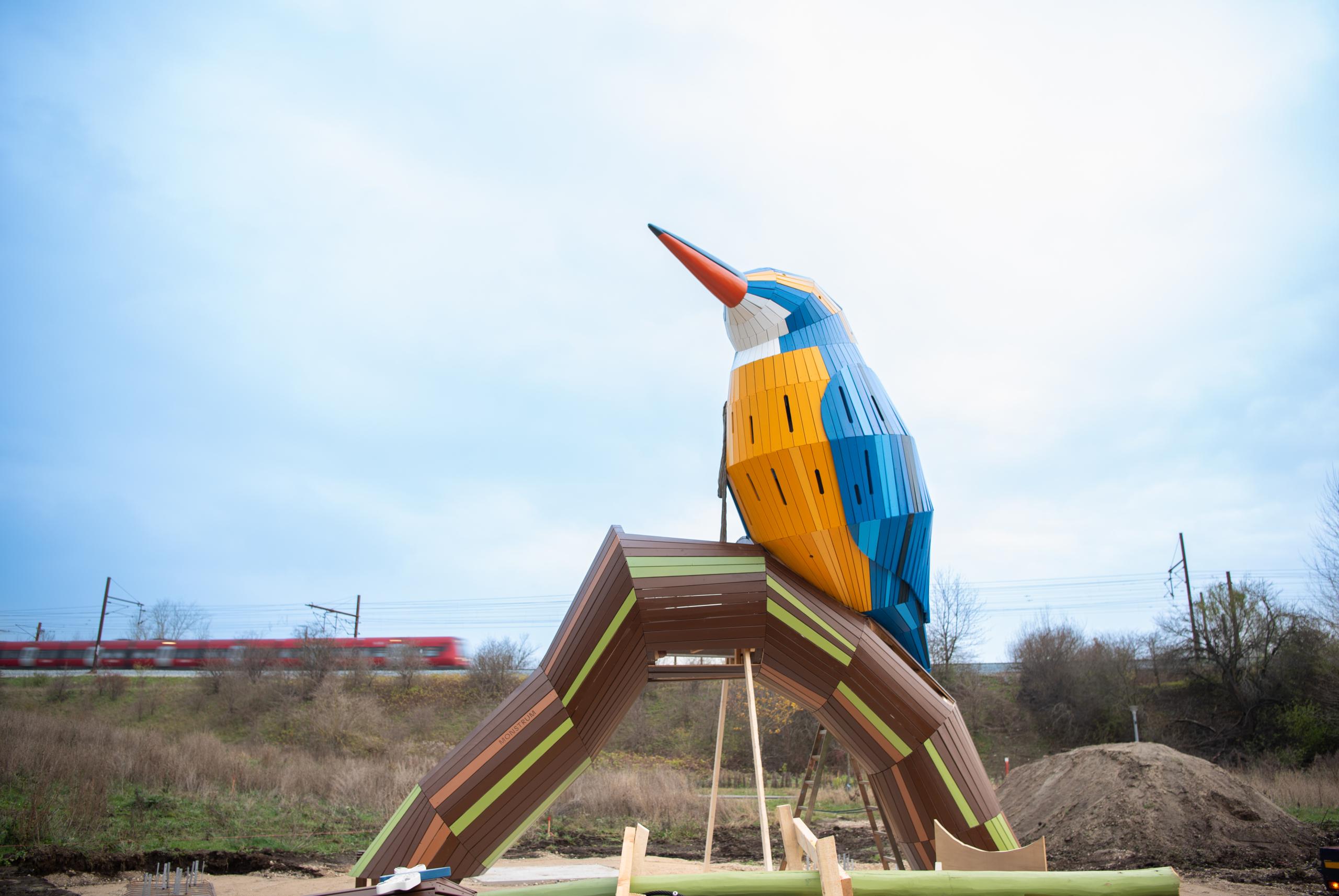 A wooden kingfisher playground structure under construction with a train passing in the background